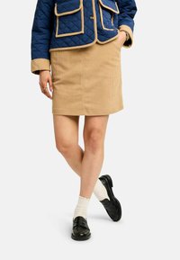 Femeie purtând o fustă bej din catifea cu caneluri, jachetă matlasată bleumarin cu margini căprui, șosete albe și mocasini negri, stând cu un picior peste altul.