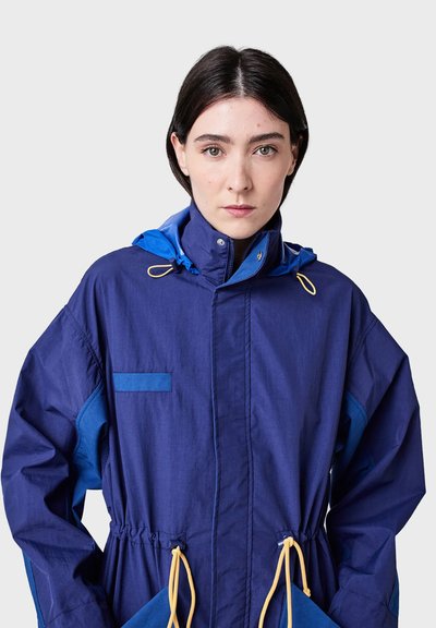 Blue outdoor jacket with a hood, elastic drawstring waist, large front pockets, contrasting blue accents, and a smooth fabric texture.