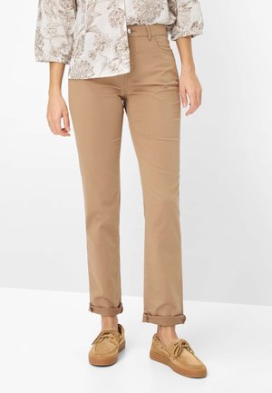 Femme portant un pantalon beige à jambes droites avec des ourlets retroussés, un haut à motif floral, et des chaussures décontractées à lacets marron clair.