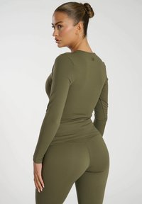 Frau mit einem Haarknoten, die ein eng anliegendes olivgrünes Langarmoberteil und Leggings trägt, steht und schaut über ihre Schulter vor einem weißen Hintergrund.
