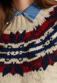Beige stickad tröja med rund hals, färgglad fair isle-mönster i röd, marinblå och vit, med en ljus denimskjorta under.