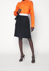 Oranssi nappikauluspaita pitkillä hihoilla, yhdistetty mustaan hameeseen, jossa on etupurut ja valkoinen tikkaus. Mustat korot sandaalit.