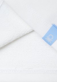 Tessuto di asciugamano bianco con superficie texture, caratterizzato da un'etichetta blu chiaro con un logo. Dettaglio della cucitura visibile sul bordo.
