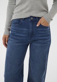 Personne portant un jean en denim bleu et un pull à manches longues gris à côtes, une main dans la poche avant et l'autre à côté de la hanche.