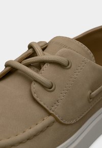 Μπεζ δερμάτινο παπούτσι τύπου boat με υφή στην επιφάνεια, στρογγυλές οπές για κορδόνια, κορδόνια στο ίδιο χρώμα, λευκή λαστιχένια σόλα και εμφανείς ραφές ως διακοσμητικά στοιχεία.