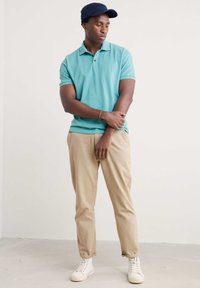 Turkos polo skjorta med korta ärmar, beiga chinos och vita sneakers. Modellen bär en marinblå keps, med korsade armar, och har en slät textur.