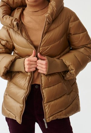 Personne portant un pull à col roulé beige et dézippant une doudoune matelassée beige par-dessus un pantalon foncé, vue du cou jusqu'aux hanches.