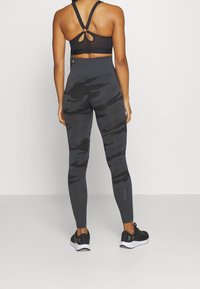 Top sportivo nero con spalline incrociate e leggings mimetici a vita alta in toni di grigio scuro. Le calzature sono nere con suole bianche.