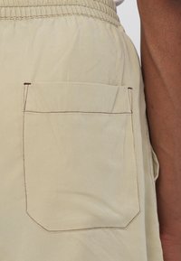 Beige Shorts mit elastischem Bund und einer quadratischen Gesäßtasche. Die Tasche ist mit kontrastierenden braunen Nähten verziert. Glattes, leichtes Material.