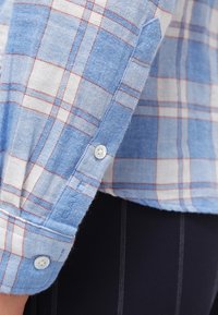 Modrá a bílá kostkovaná košile s červenými detaily, zapínáním na knoflíky a ohrnutými manžetami. Vyrobena z měkké látky s hladkým povrchem.