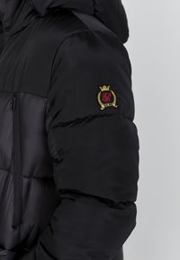 Schwarze Pufferjacke mit einem gesteppten Muster, ausgestattet mit einem Reißverschluss und einem gestickten Logo in Gold, Rot und Schwarz auf der linken Schulter.