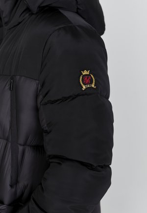 Czarna kurtka puchowa z zapięciem na zamek, pikowaną teksturą oraz haftowanym złotym i czerwonym logo na lewym rękawie.