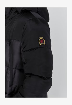 Must musta jakk, millel on lukuga sulgemine, tikitud tekstuur ja kuldne ning punane brodeeritud logo vasakul varrukal.