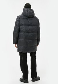 Lang, mørkegrå dunjakke med hætte, der har stitched vandrette sektioner, blød tekstur og en afslappet pasform. Parret med sorte bukser.