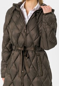 Quiltad pufferjacka i mörk olivgrön, med en åtstramande midja med bälte, huva och sidofickor. Slätt, lätt tyg.