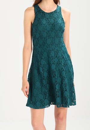 Bezrekawna sukienka z tiulu w kolorze morskim, dopasowany gorset i rozkloszowany dół, noszona przez stojącą osobę na jednolitym tle.