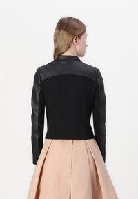 Giacca nera aderente con maniche in pelle e corpo in tessuto liscio. La parte posteriore presenta dettagli delle cuciture e un colletto appuntito.