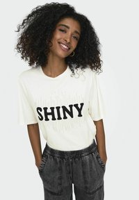 Cremefarvet T-shirt med fed sort "SHINY" tekst og rhinsten-detaljer, parret med afslappede mørkegrå bukser. Blødt stof, korte ærmer.