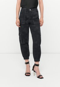 Zwarte camouflage cargo pantalons met elastische boorden, twee voorzakken en een knoopsluiting. Gecombineerd met zwarte sandalen met hoge hakken en banden.