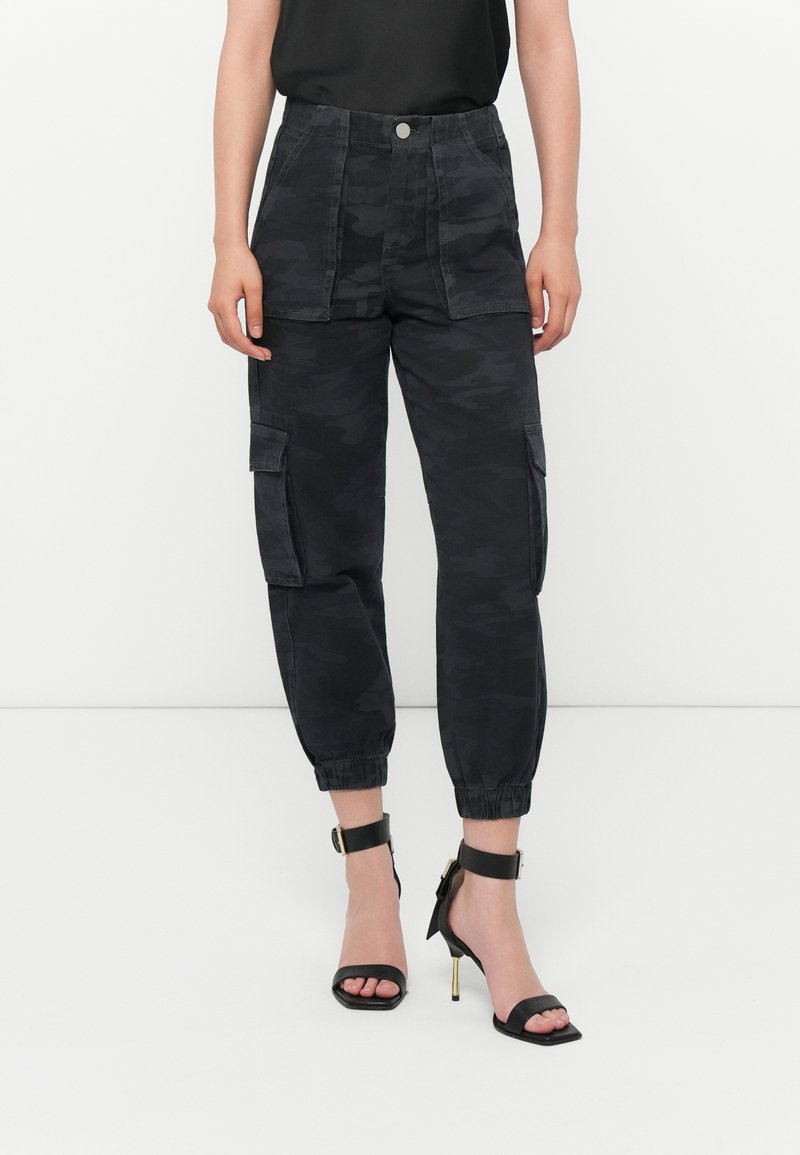Zwarte camouflage cargo pantalons met elastische boorden, twee voorzakken en een knoopsluiting. Gecombineerd met zwarte sandalen met hoge hakken en banden.