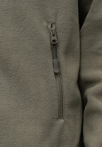 Κοντινό πλάνο μιας φλις ζακέτας ελιάς με κάθετη τσέπη με φερμουάρ και υφή υφάσματος.