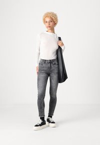 Haut à manches longues blanc, jeans gris ajustés, baskets noires avec semelles blanches, et un sac noir, sur fond uni.