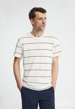 Mann mit kurzen braunen Haaren, der ein weißes T-Shirt mit dünnen schwarzen horizontalen Streifen und marineblaue Hosen trägt, die Hände in den Taschen hat und nach rechts schaut.