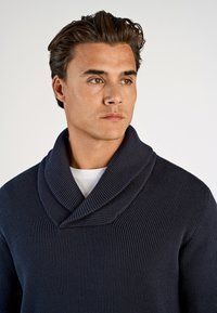 Marineblaue Rippstrickpullover mit weiter Schalkragen, aus strukturiertem Material und schlanker Passform. Über einem weißen T-Shirt getragen.