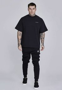 Schwarzes T-Shirt mit kurzen Ärmeln und schwarze Jogginghosen, beide mit lockerer Passform. Das T-Shirt hat ein kleines Logo auf der Brust; die Jogginghosen sind mit einem weißen Akzent versehen.