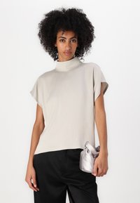Top beige chiaro a collo alto con maniche corte, tessuto morbido, vestibilità ampia, abbinato a una piccola borsa a tracolla in argento e pantaloni neri a gamba larga.