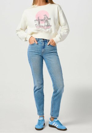 Femme portant un sweat-shirt écru avec un motif de cowboy et un jean bleu clair, debout les mains dans les poches, avec des baskets bleues.