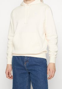 Ljust persikofärgad hoodie av mjukt, texturerat tyg med en kenguruficka och en vit Nike-logotyp på vänster bröst. Kombinerad med blå jeans.