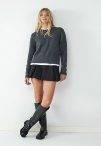 Grauer Pullover mit Rundhalsausschnitt, über einem weißen Hemd. Schwarzer, ausgestellter Minirock und knielange schwarze Stiefel mit blockiertem Absatz.