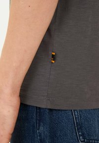 Grå kortærmet skjorte med en tekstureret overflade, med et sideetiket i sort og orange detaljer, parret med blå denim.
