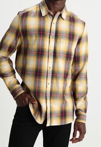 Camicia di flanella a quadri gialla con accenti rossi e neri, colletto button-down e maniche lunghe. Tessuto morbido e vestibilità rilassata.