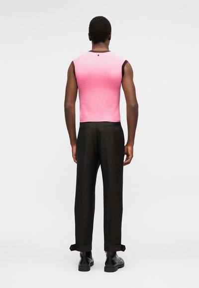 Άνδρας με το βλέμμα προς τα πίσω, φοράει ροζ αμάνικη μπλούζα ombre, μαύρο παντελόνι με κουμπωτά μανίκια και μαύρα δερμάτινα μποτάκια σε λευκό φόντο.