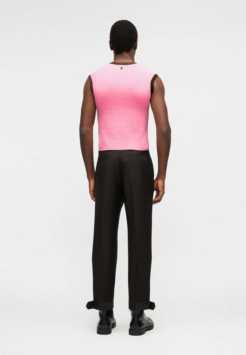 Man facing away wearing sleeveless pink ombre top, black tailored pants with buttoned cuffs, and black leather boots on a white background.