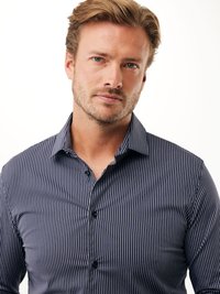 Uomo biondo con una leggera barba, che indossa una camicia a righe blu scuro e bianca con bottoni, mentre guarda leggermente verso la fotocamera.