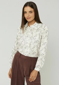 Chemisier blanc avec imprimé floral, col froncé et boutonnage à l'avant. Manches longues et transparentes avec une texture en couches, associé à un pantalon marron taille haute.