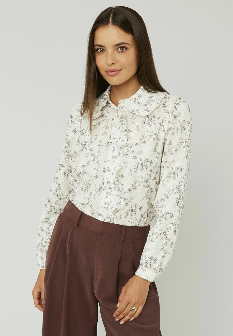 Chemisier blanc avec imprimé floral, col froncé et boutonnage à l'avant. Manches longues et transparentes avec une texture en couches, associé à un pantalon marron taille haute.