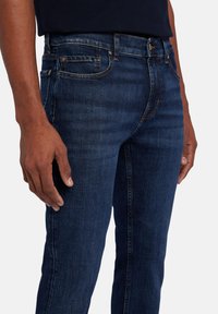 Jean en denim bleu foncé avec une coupe slim, présentant des coutures visibles, un design à cinq poches et une taille mi-haute. Le tissu a une texture lisse.