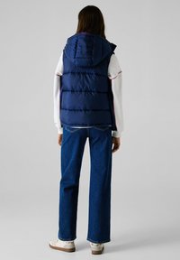 Ναυτικό μπλε γιλέκο πούφ με κουκούλα, με καπιτονέ σχέδιο. Φοριέται πάνω από ένα λευκό μακρυμάνικο πουκάμισο και συνδυάζεται με παντελόνι φαρδύ.