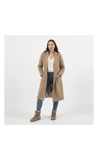 Long manteau beige avec un col, deux poches avant, associé à un pull blanc texturé et un jean bleu, complété par des bottines tan.