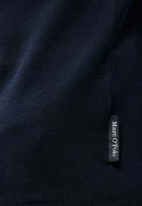 Ναυτικό μπλε ύφασμα με λεία υφή, που διαθέτει μία μικρή μαύρη ετικέτα με λευκό κείμενο που αναγράφει "Marc O'Polo" κοντά στην άκρη.