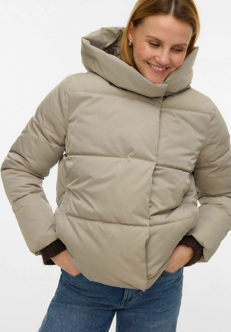 Veste matelassée beige avec une grande capuche, présentant un design matelassé et une coupe carrée. Le tissu est lisse avec un léger éclat.