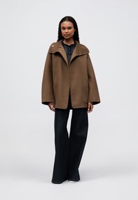 Abrigo de mezcla de lana marrón con un amplio cuello, dos bolsillos frontales y un detalle de un solo botón, combinado con jeans de mezclilla oscuros acampanados.