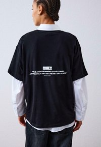 Čierne oversized tričko s krátkymi rukávmi, na zadnej strane s citátom v bielej farbe, prekryté bielou košou.