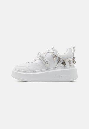Biały sneaker na platformie z grubą podeszwą, paskiem z nazwą marki, bocznym zamkiem błyskawicznym oraz srebrnym łańcuszkiem z zawieszkami owiniętym wokół pięty.