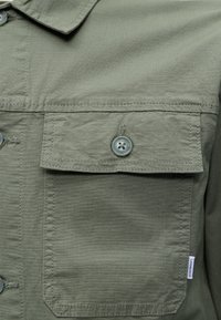 Poche de chemise en tissu texturé vert olive avec rabat boutonné et petite étiquette blanche de la marque sur le côté droit.
