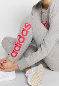 Pantaloni jogger grigi in cotone con il logo "adidas" in bold rosa lungo la gamba, abbinati a scarpe da ginnastica bianche con suola texturizzata.